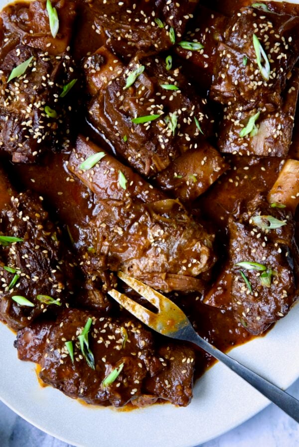Korean Braised Beef Short Ribs on oval white platter with meat fork sprinkled with sesame seeds and thinly sliced scallions.