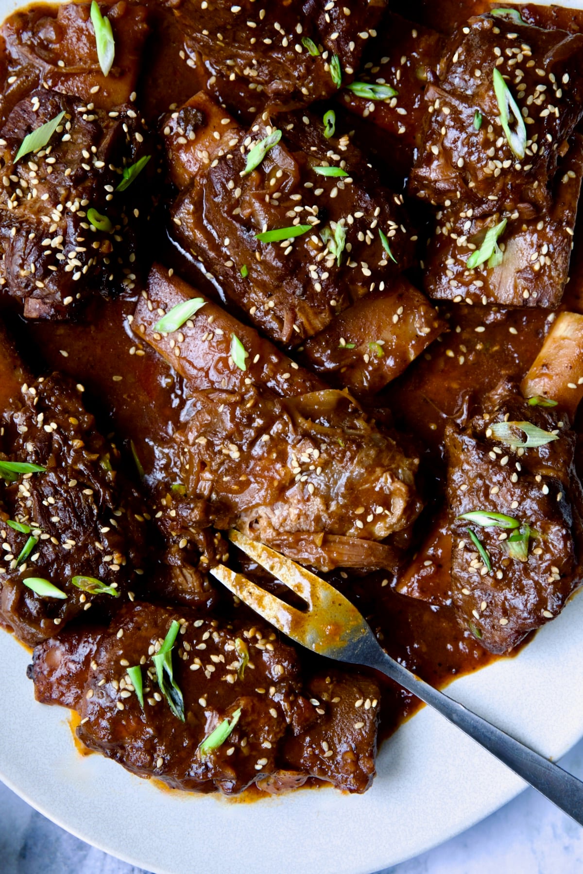 Korean Braised Beef Short Ribs on oval white platter with meat fork sprinkled with sesame seeds and thinly sliced scallions.