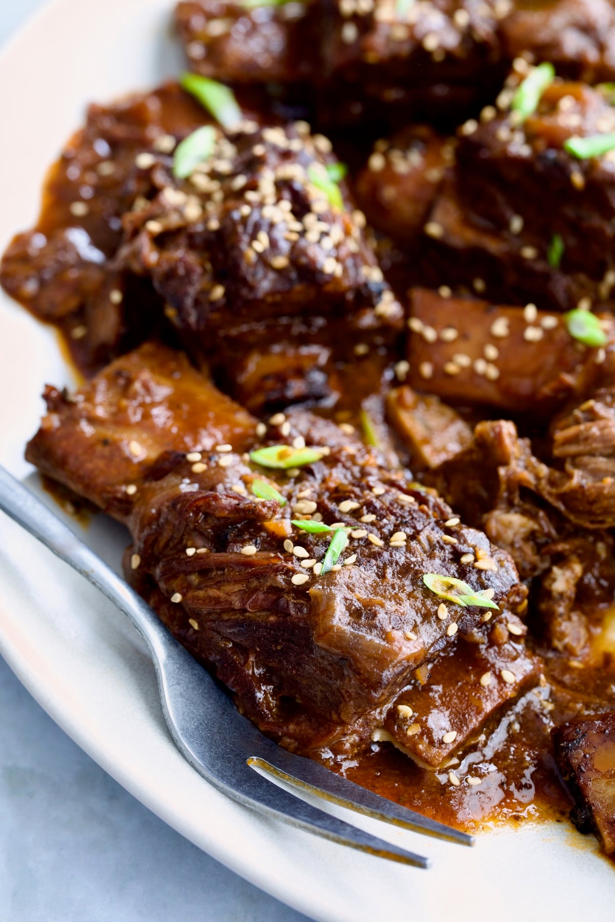 Close-up of one Korean Braised Beef Short Rib on white platter with meat fork sprinkled with sesame seeds and thinly sliced scallions.