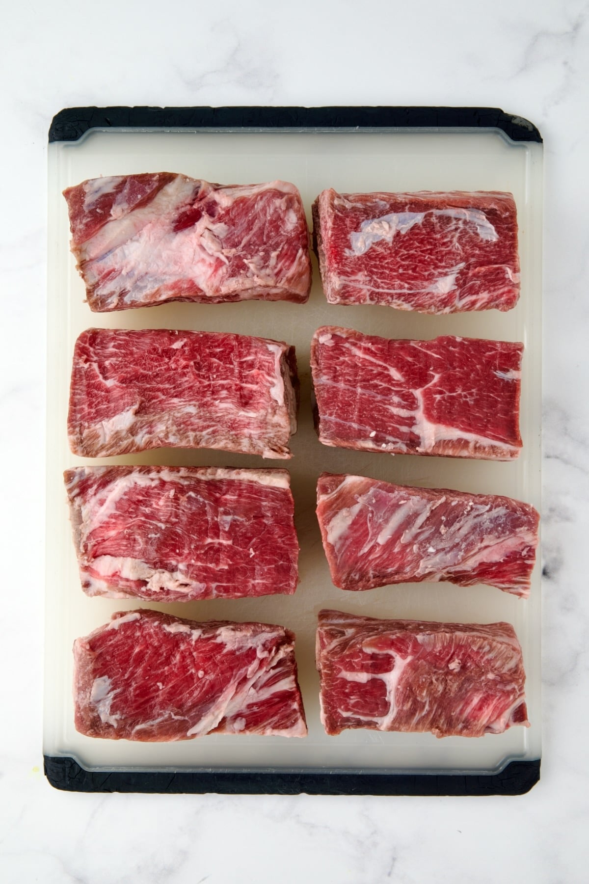 Four uncooked beef short ribs on white plastic cutting board.