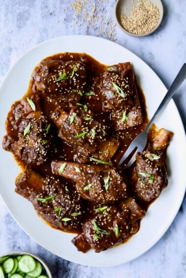 Korean Braised Beef Short Ribs on oval white platter with meat fork sprinkled with sesame seeds and thinly sliced scallions.