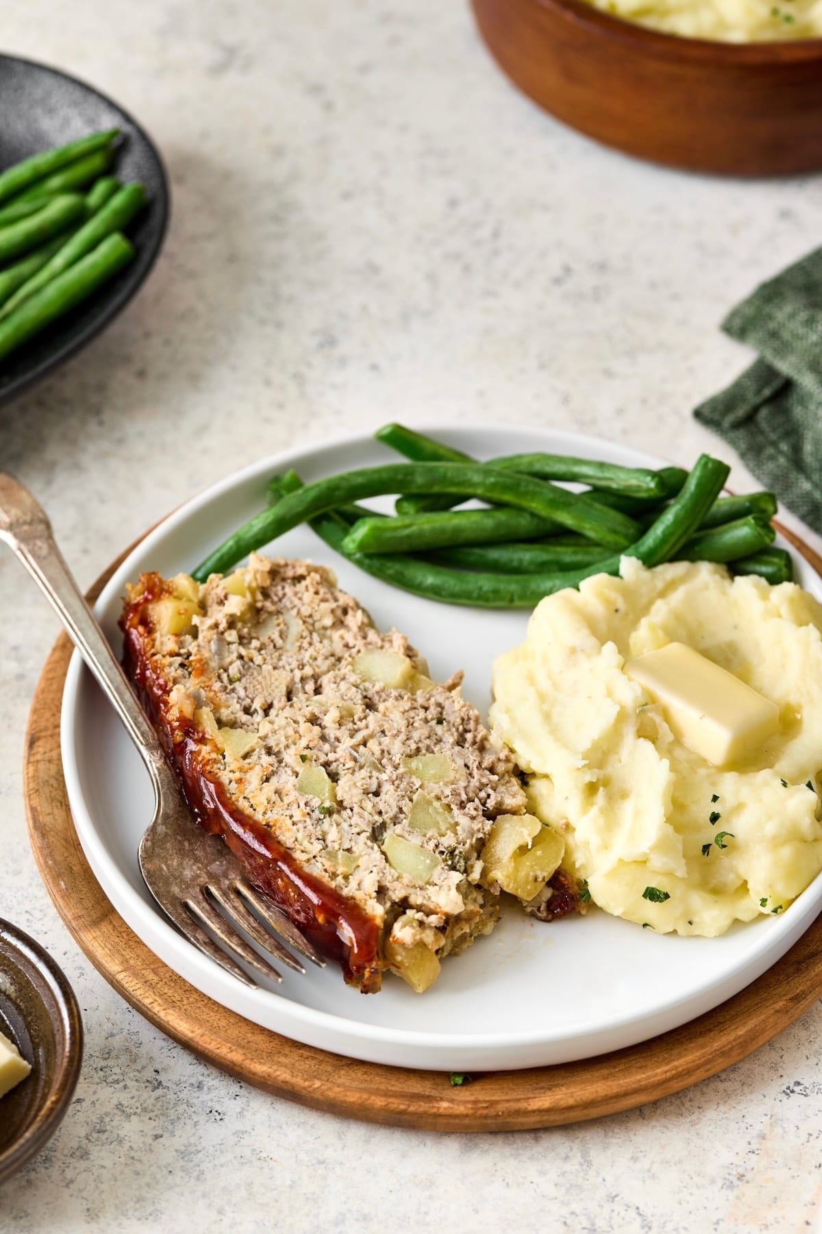 Slice of meatloaf, mashed potatoes and green beans on white plate with dinner fork.