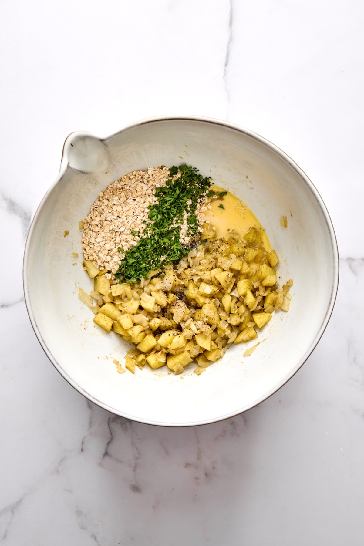 Cooked onions, apples, oatmeal, parsley and egg in white mixing bowl.