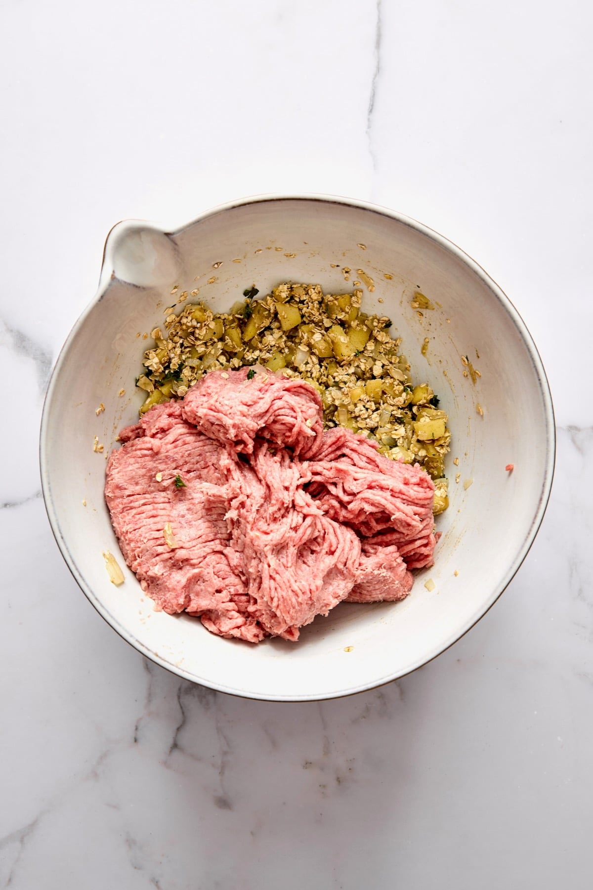 Ground turkey added to cooked onions, apples, oatmeal, parsley and egg in white mixing bowl.
