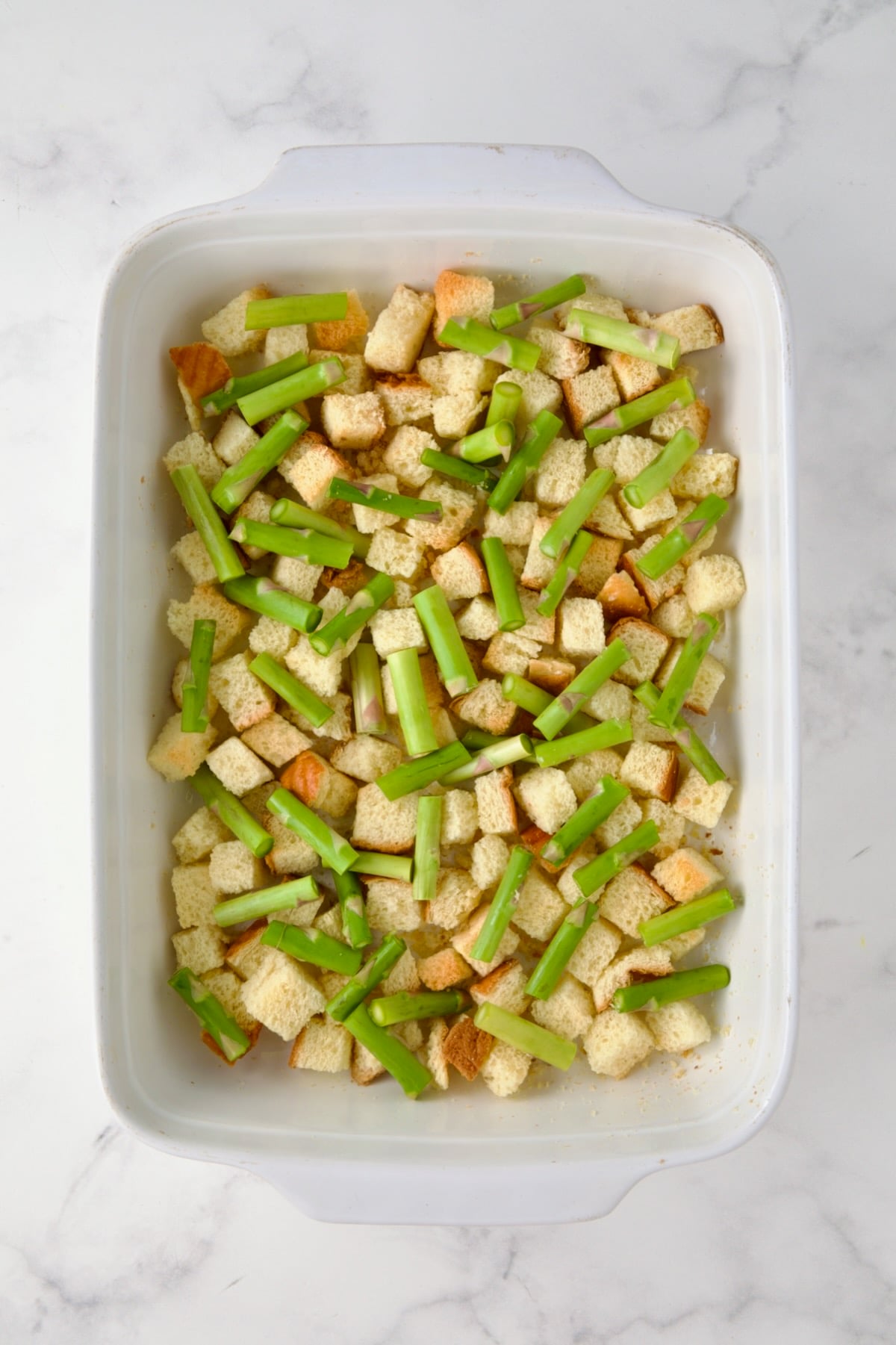 Half of the asparagus pieces over the brioche bread cubes in white rectangular baking dish.