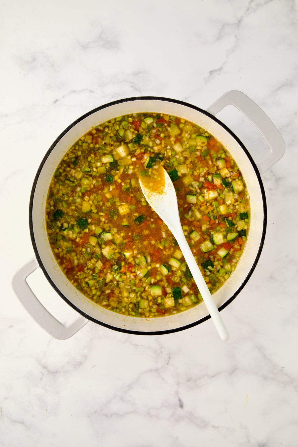 Chicken broth added to vegetables and spices in white enamel cast iron skillet.