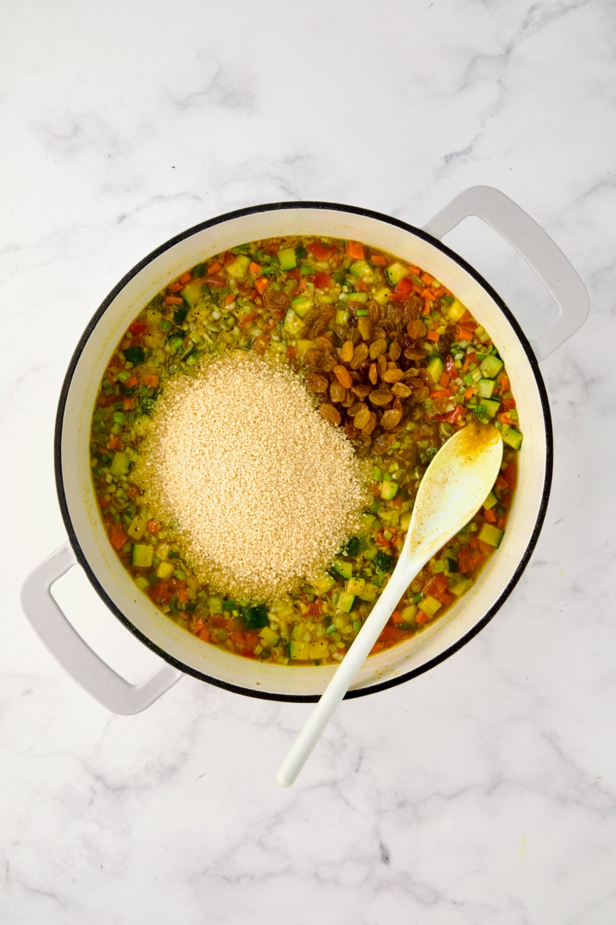 Couscous and golden raisins added to chicken broth and vegetables in white enamel cast iron skillet.