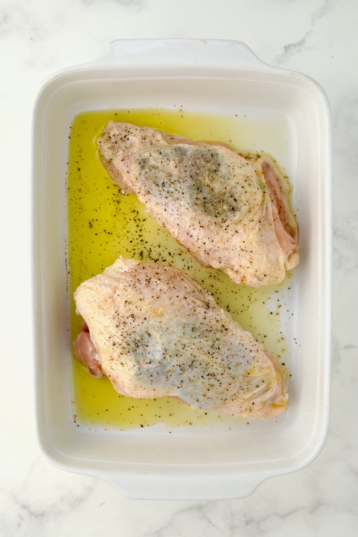 Two uncooked split chicken breasts seasoned with salt and pepper in white rectangular baking dish.