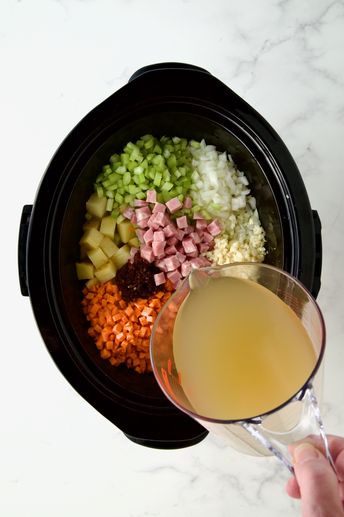Diced vegetables and ham in black slow cooker before being mixed together with chicken broth being poured in with glass pitcher.