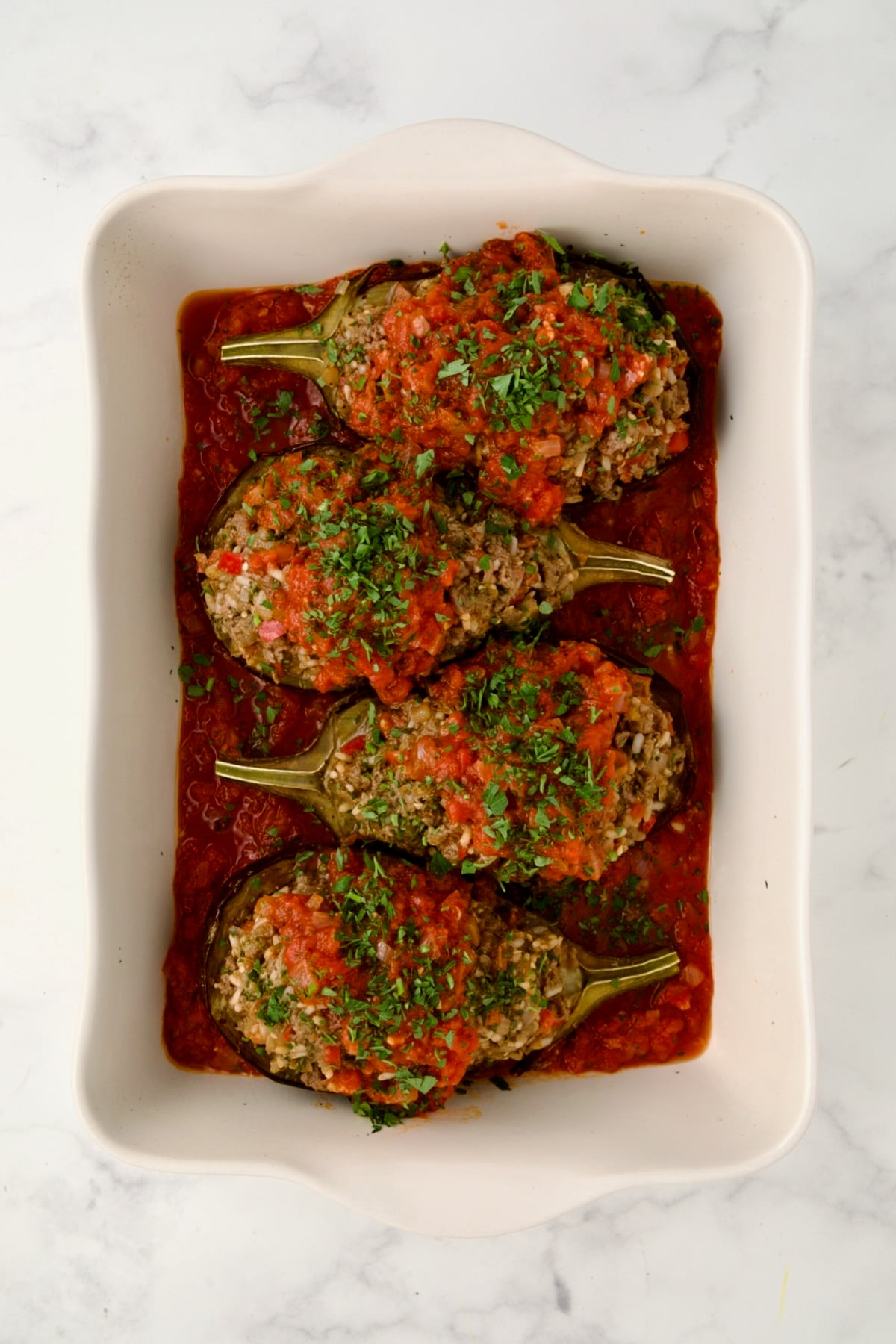 Four Stuffed Eggplant halves set a top tomato sauce in white rectangular baking dish topped with tomato sauce ready to be baked.