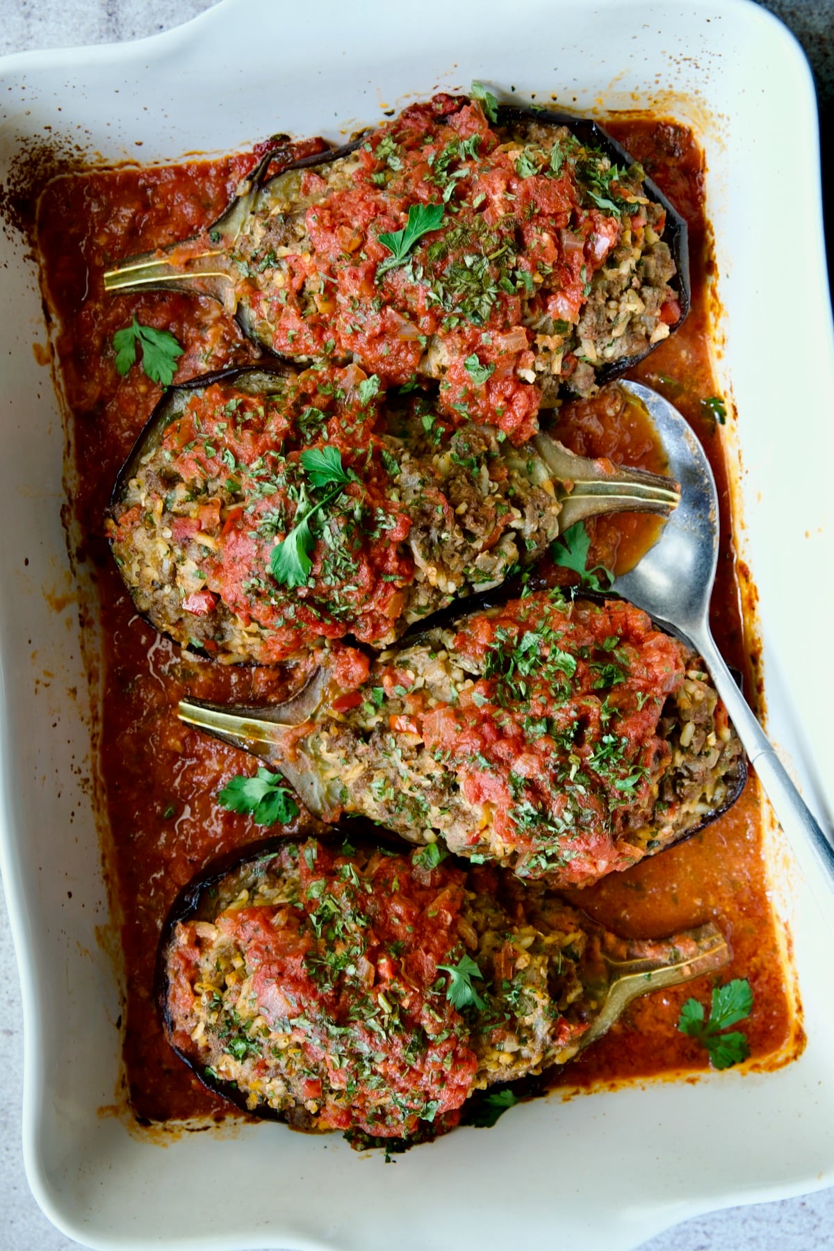 Four baked Stuffed Eggplant halves set a top tomato sauce in white rectangular baking dish with serving spoon.