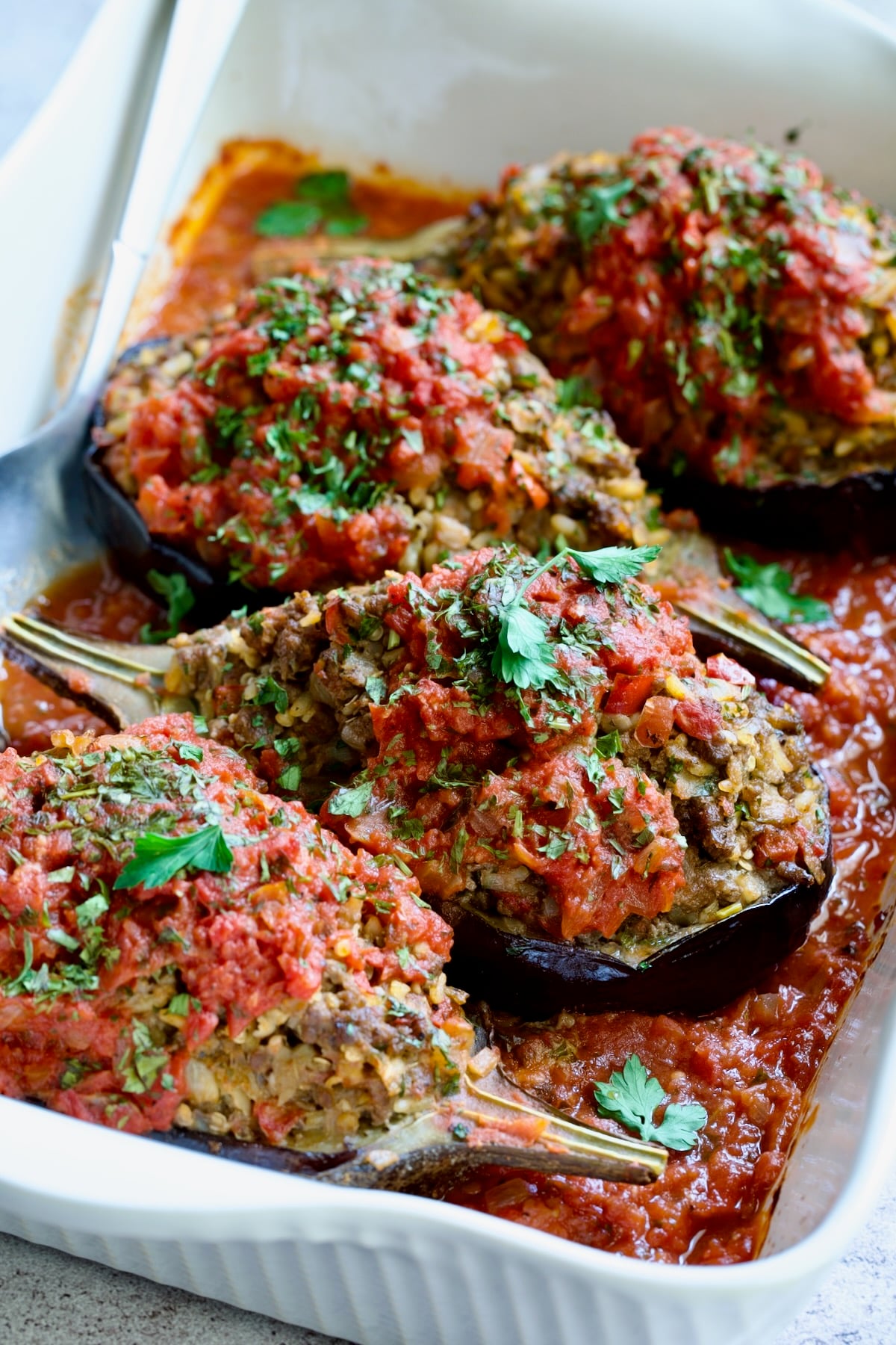 Four baked Stuffed Eggplant halves in white rectangular baking dish with serving spoon.