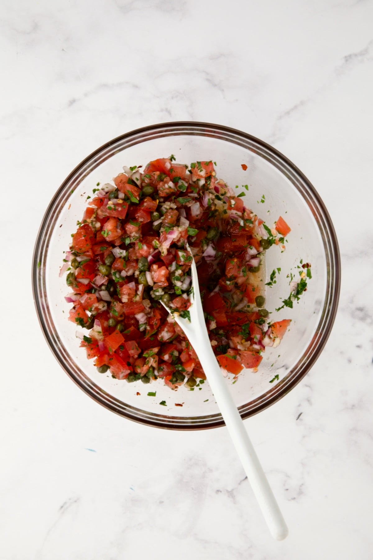 Tomato Caper Relish in glass bowl being mixed with white spoon.