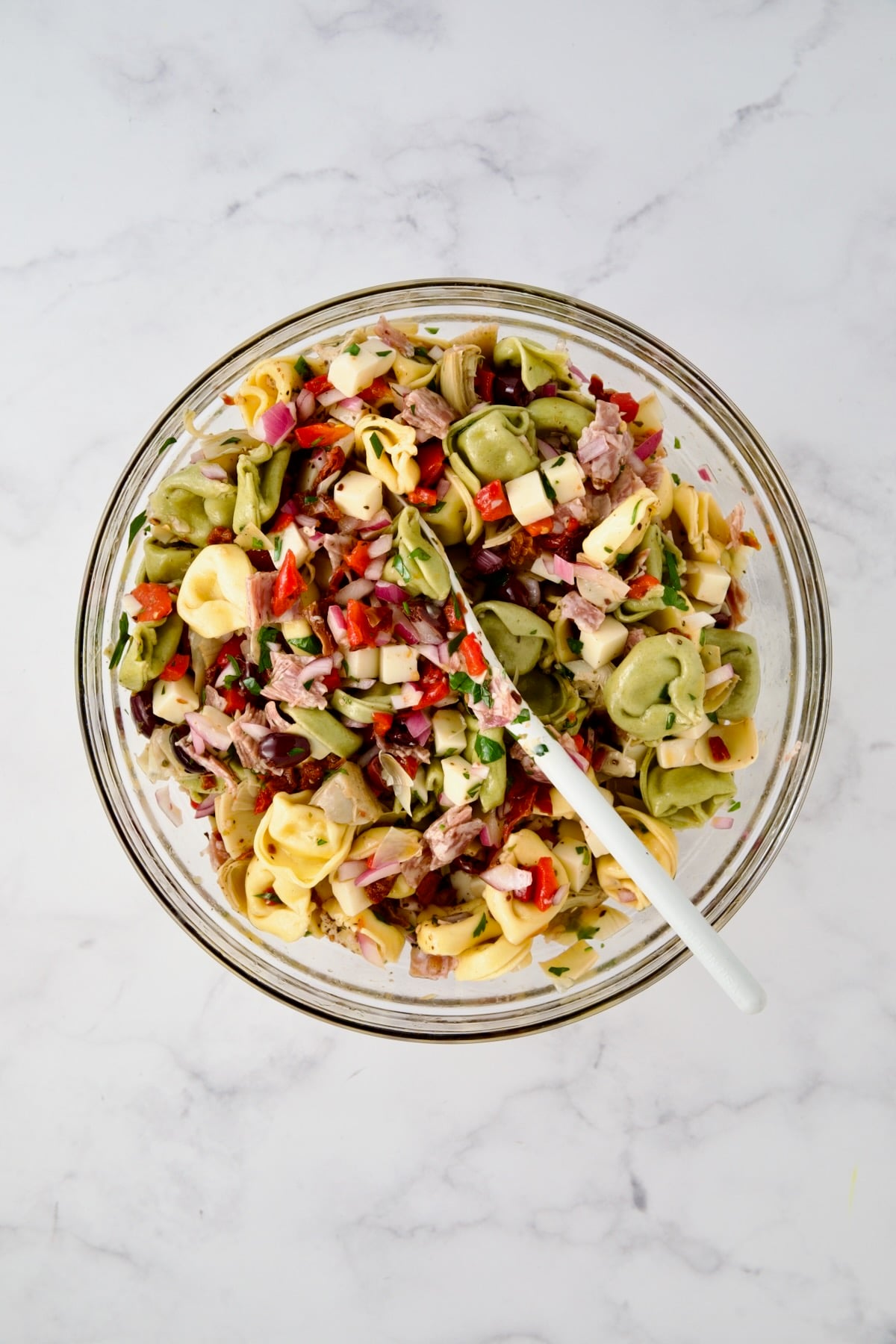 Ingredients for tortellini salad in glass mixing bowl after being combined with white mixing spoon.