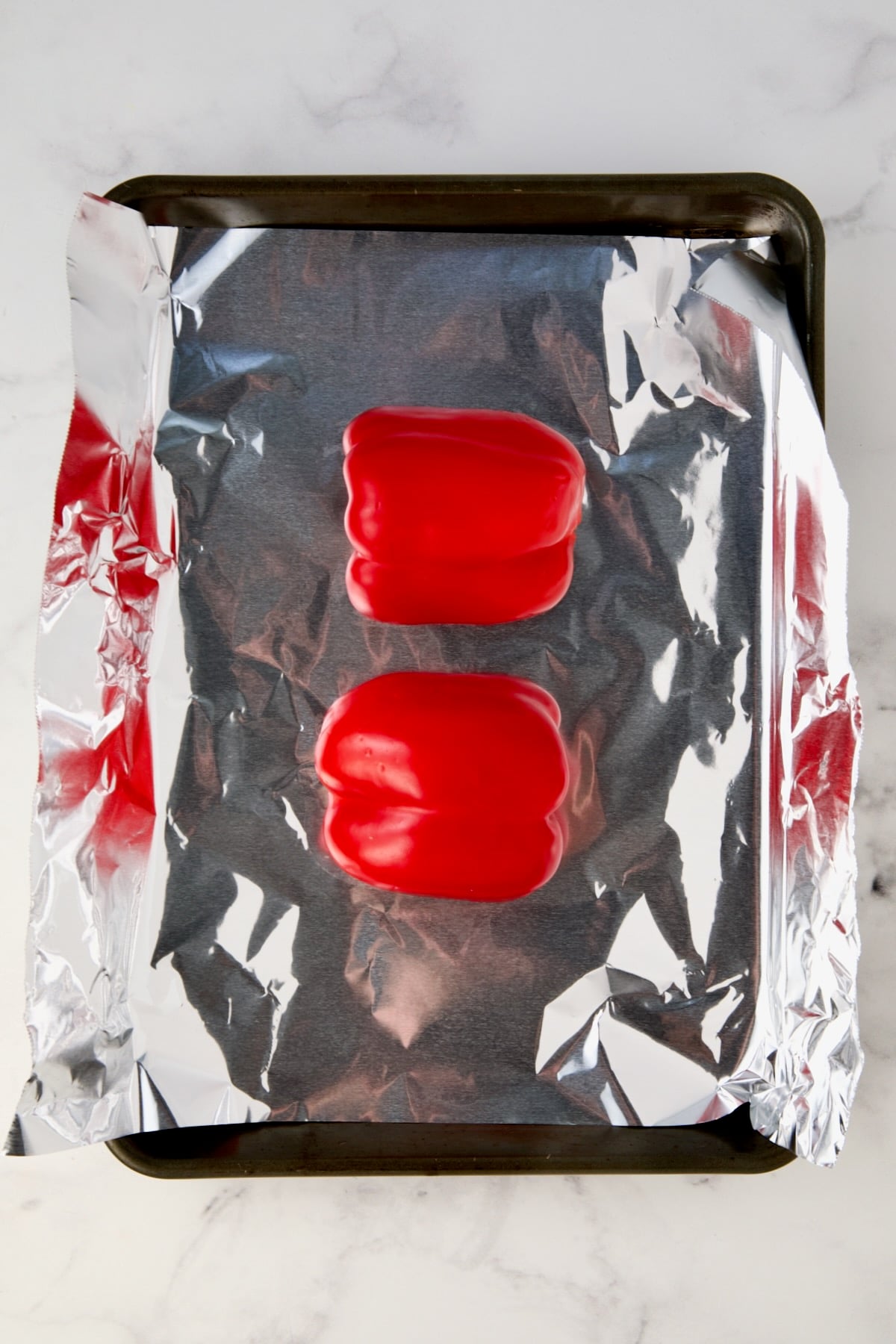 Two red bell pepper halves on aluminum foil-lined rimmed sheet pan before being roasted.