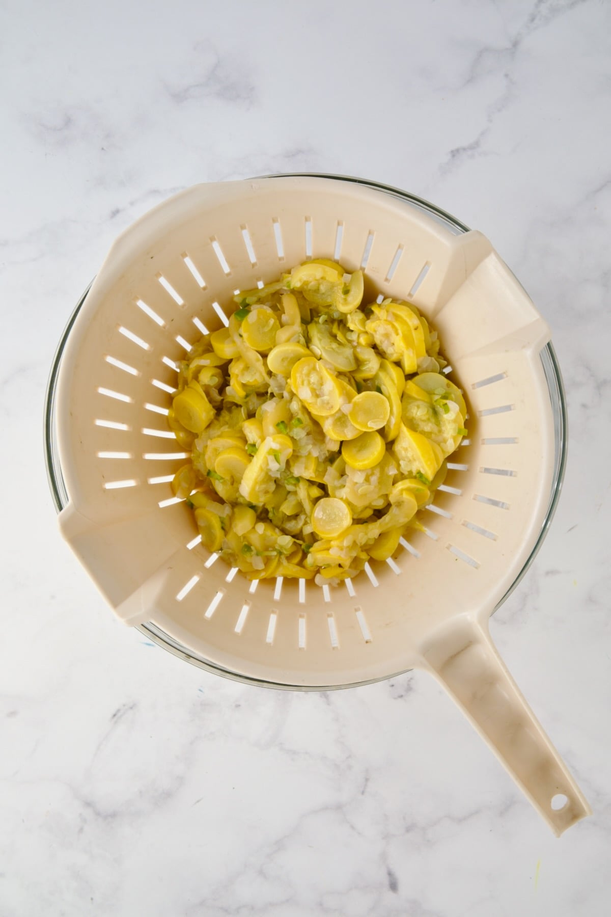 Cooked yellow squash, onion and jalapeno in beige colander set over a glass bowl.