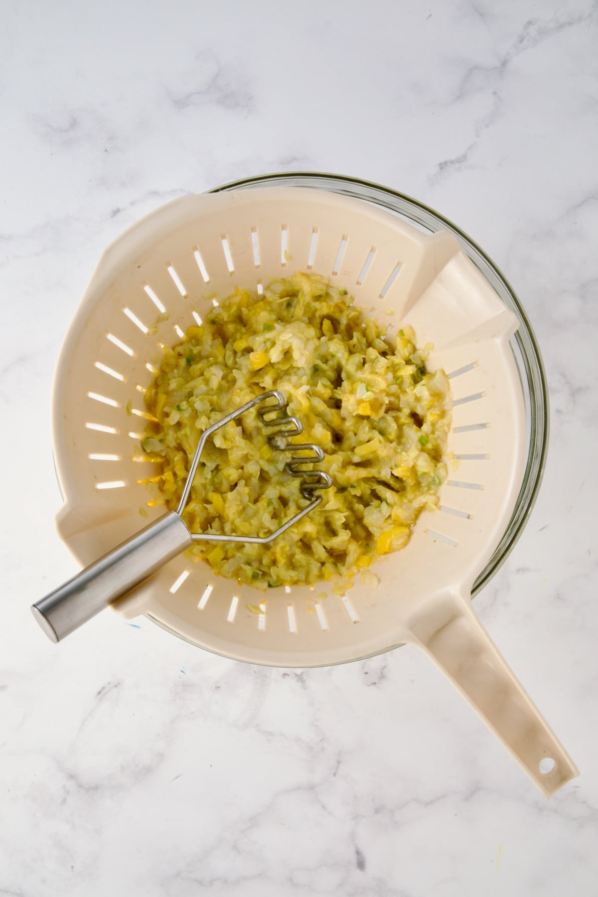 Cooked mashed yellow squash, onion and jalapeno in beige colander set over a glass bowl with potato masher.