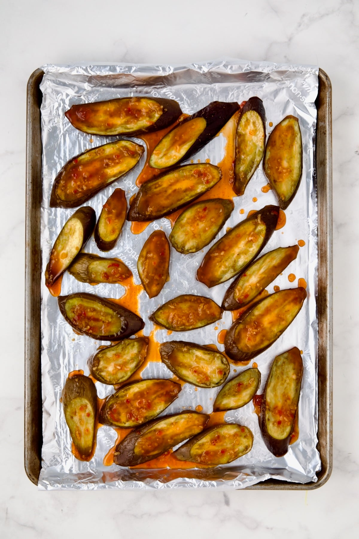 Sliced eggplant on aluminum-foil lined rimmed sheet pan after being baked with sauce drizzled over.