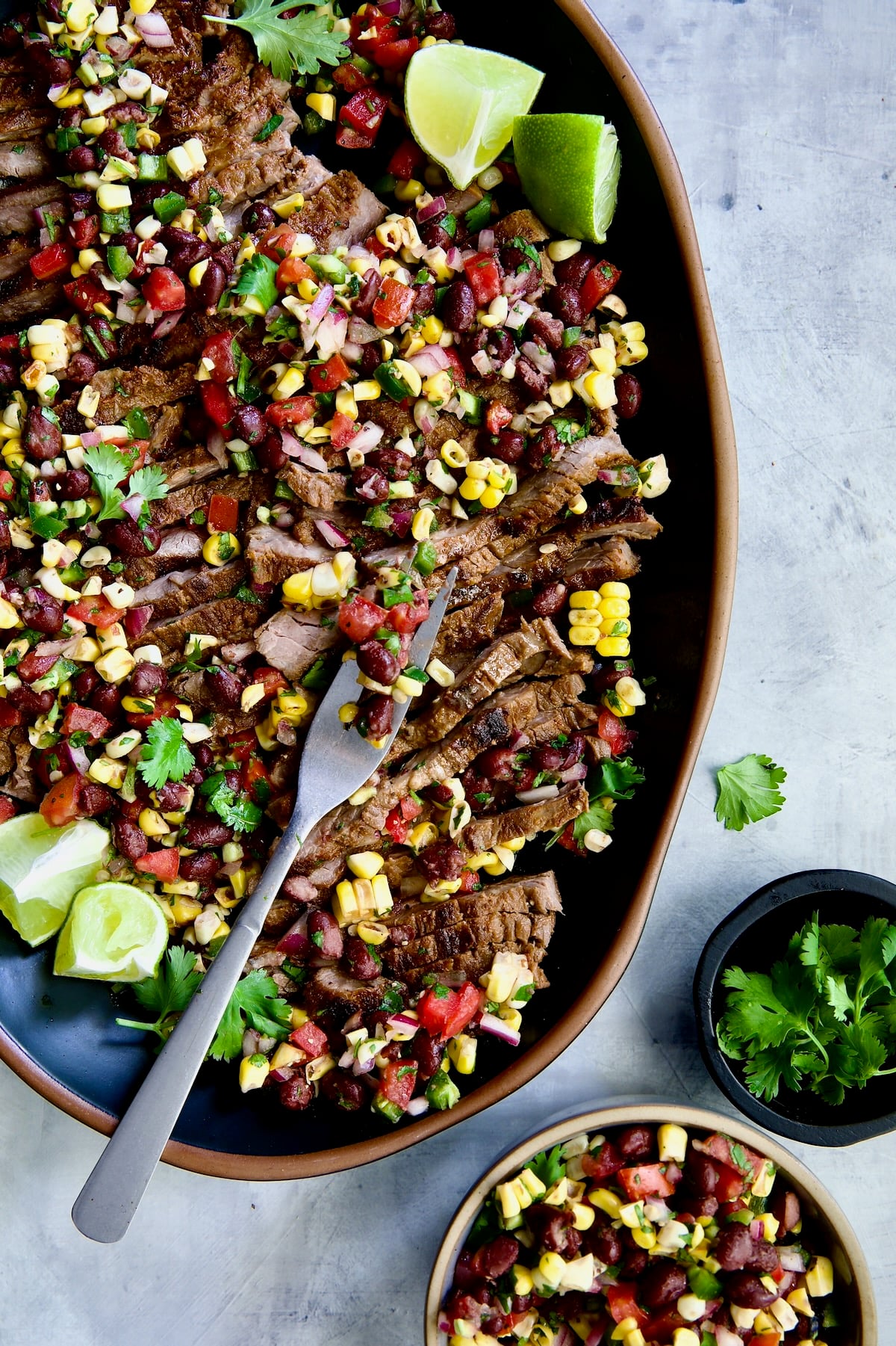 Grilled Skirt Steak with Black Bean Corn Salad on black oval platter with meat fork.