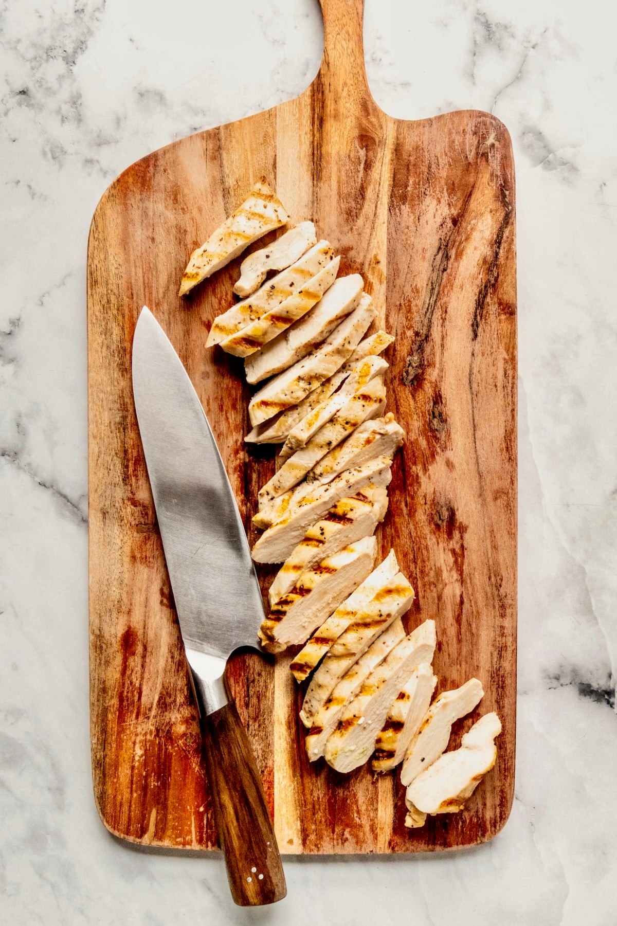 Sliced cooked chicken on wooden cutting board with chef's knife.