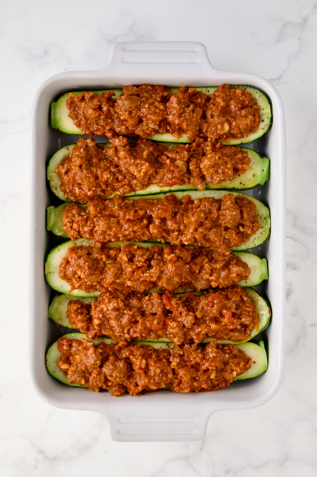 Six Chorizo-Stuffed Zucchini in white rectangular baking dish before being baked and before cheese is added.