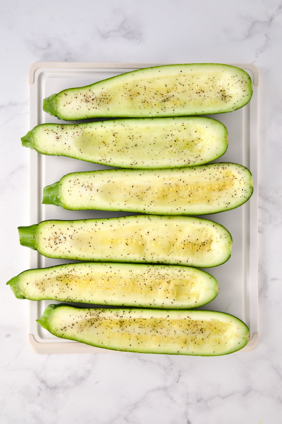 Six hollowed out zucchini halves on white plastic cutting board seasoned with salt and black pepper.