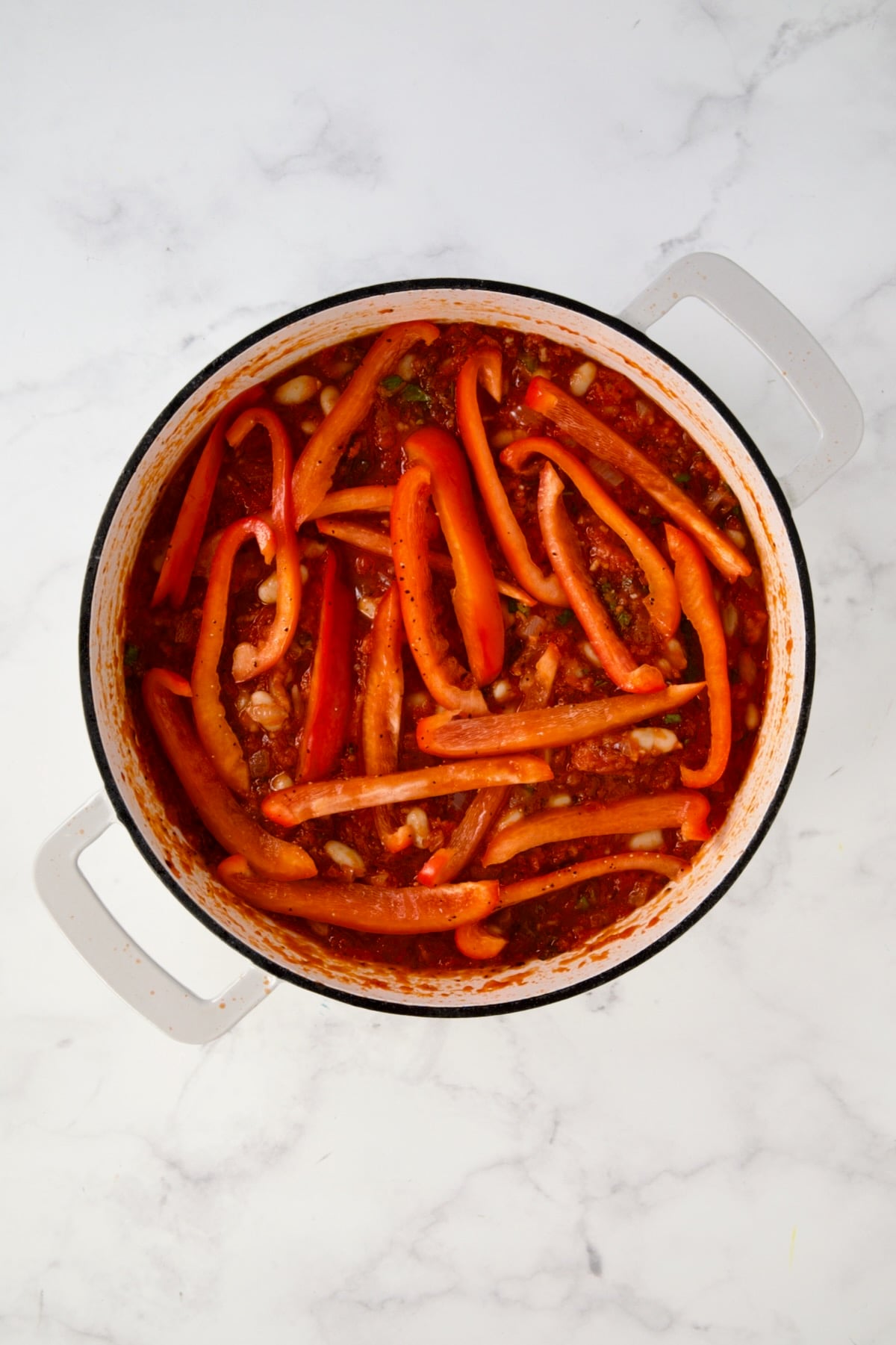 Red bell pepper slices placed over the top of tomato sauce with beans in white enamel cast iron skillet.