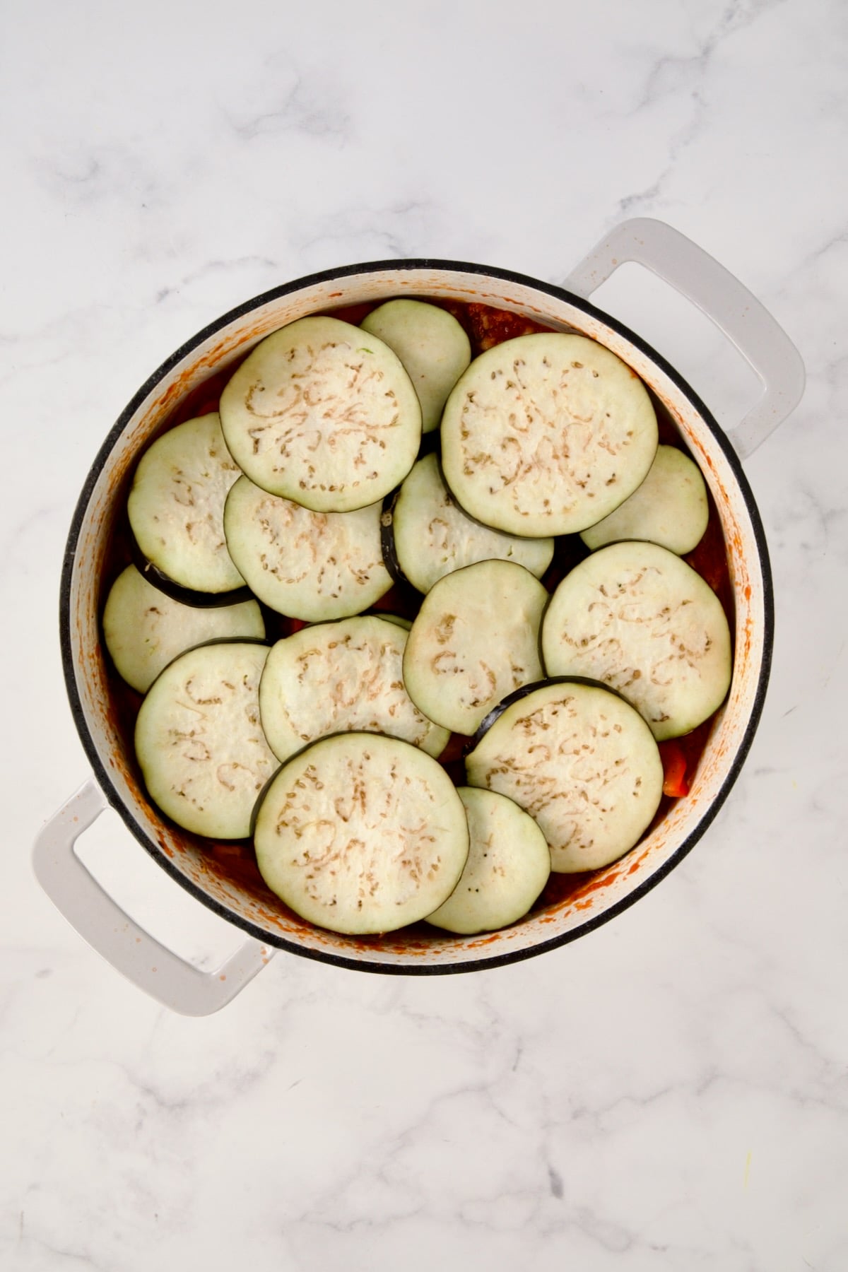 Eggplant slices placed over red bell pepper in white enamel cast iron skillet.