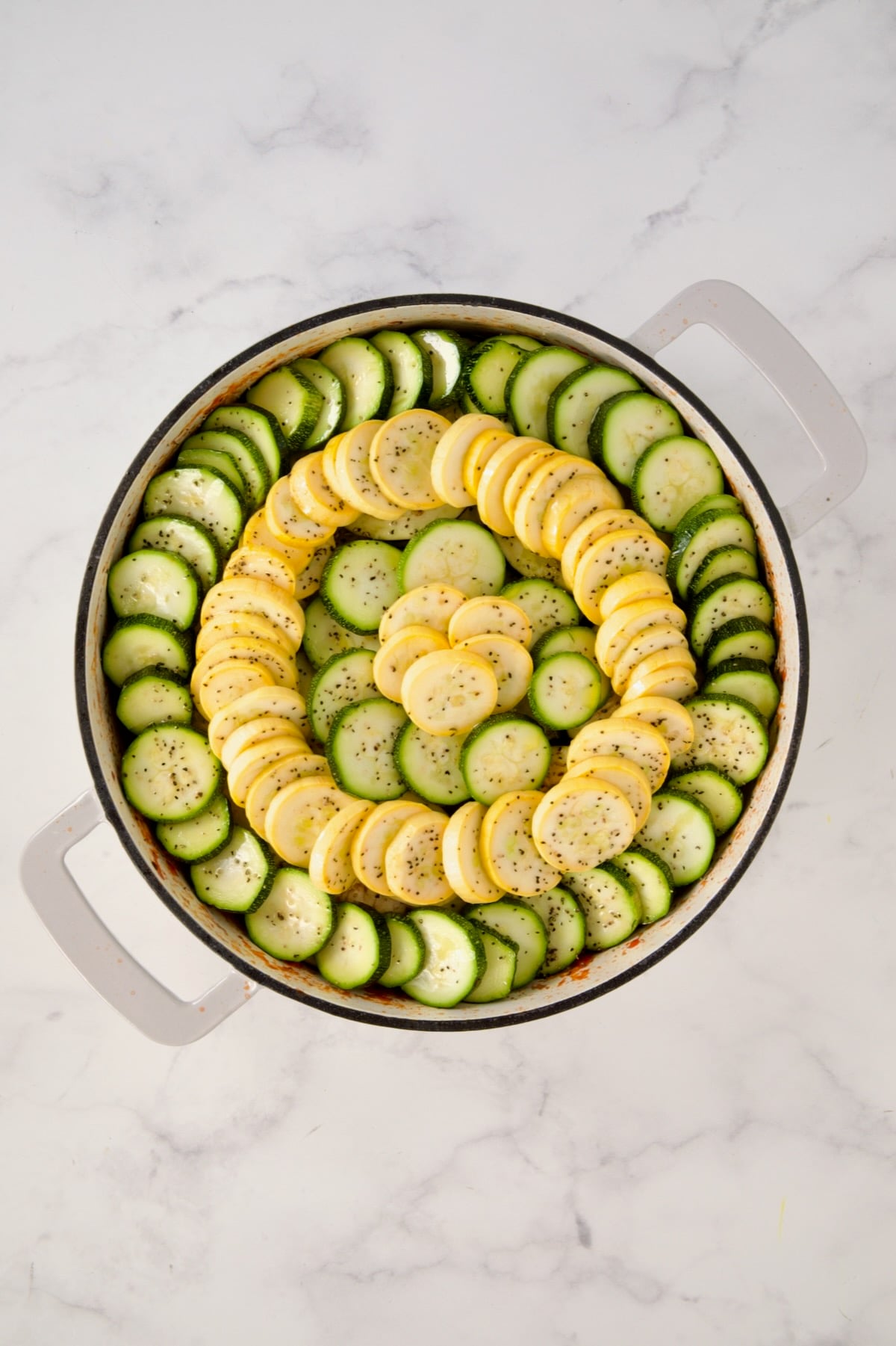 Zucchini and yellow squash slices placed over eggplant in a circular pattern in white enamel cast iron skillet.