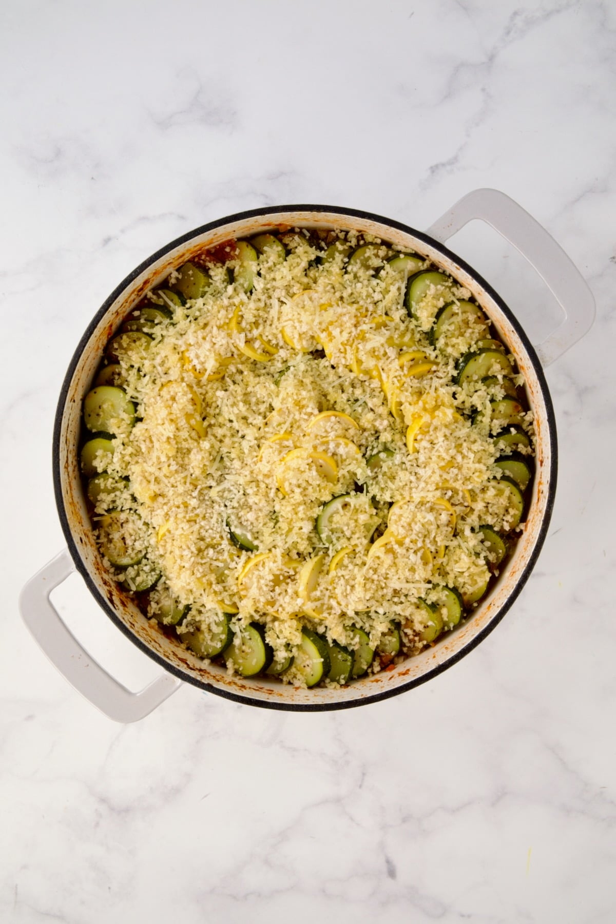 Breadcrumb mixture placed over zucchini and yellow squash in white enamel cast iron skillet.