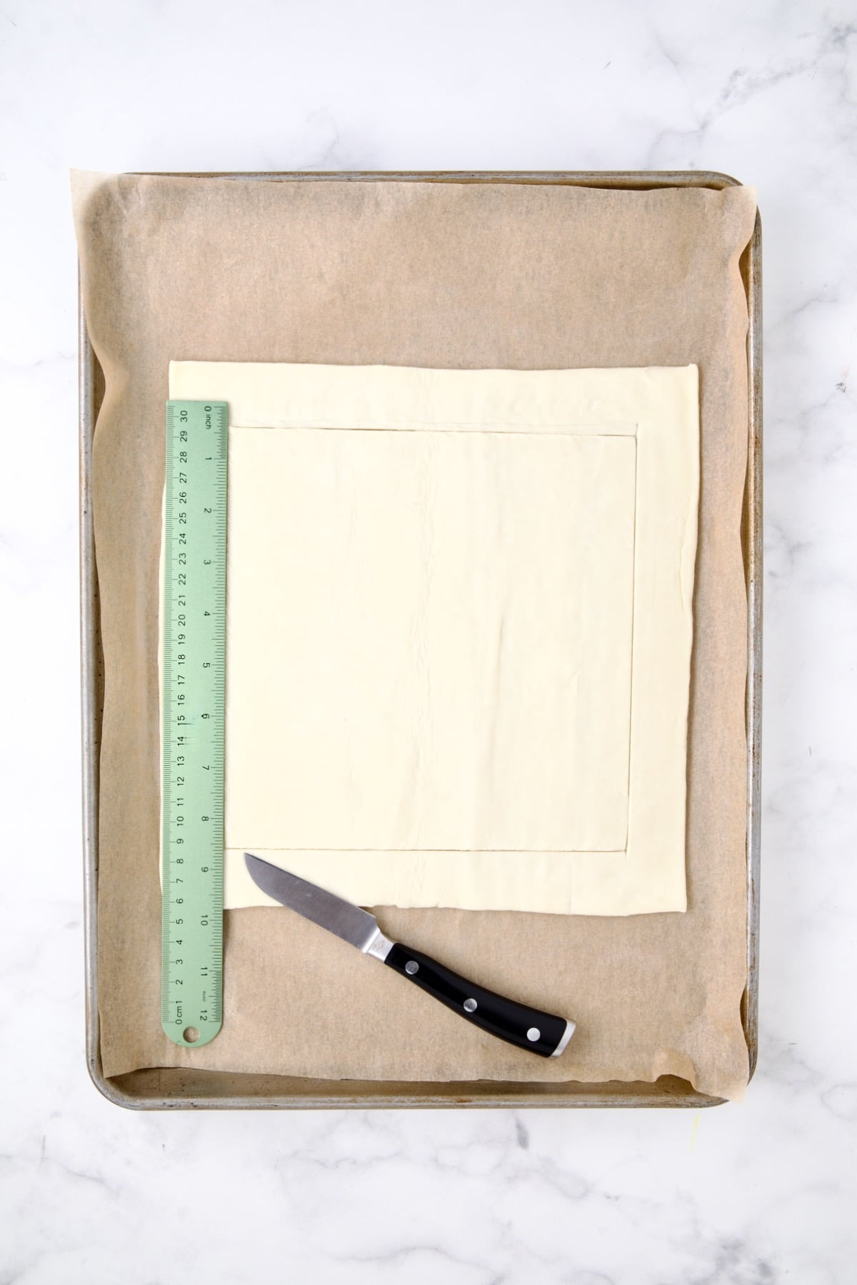 Square of puff pastry on parchment paper-lined rimmed baking sheet with knife and ruler scoring the pastry.