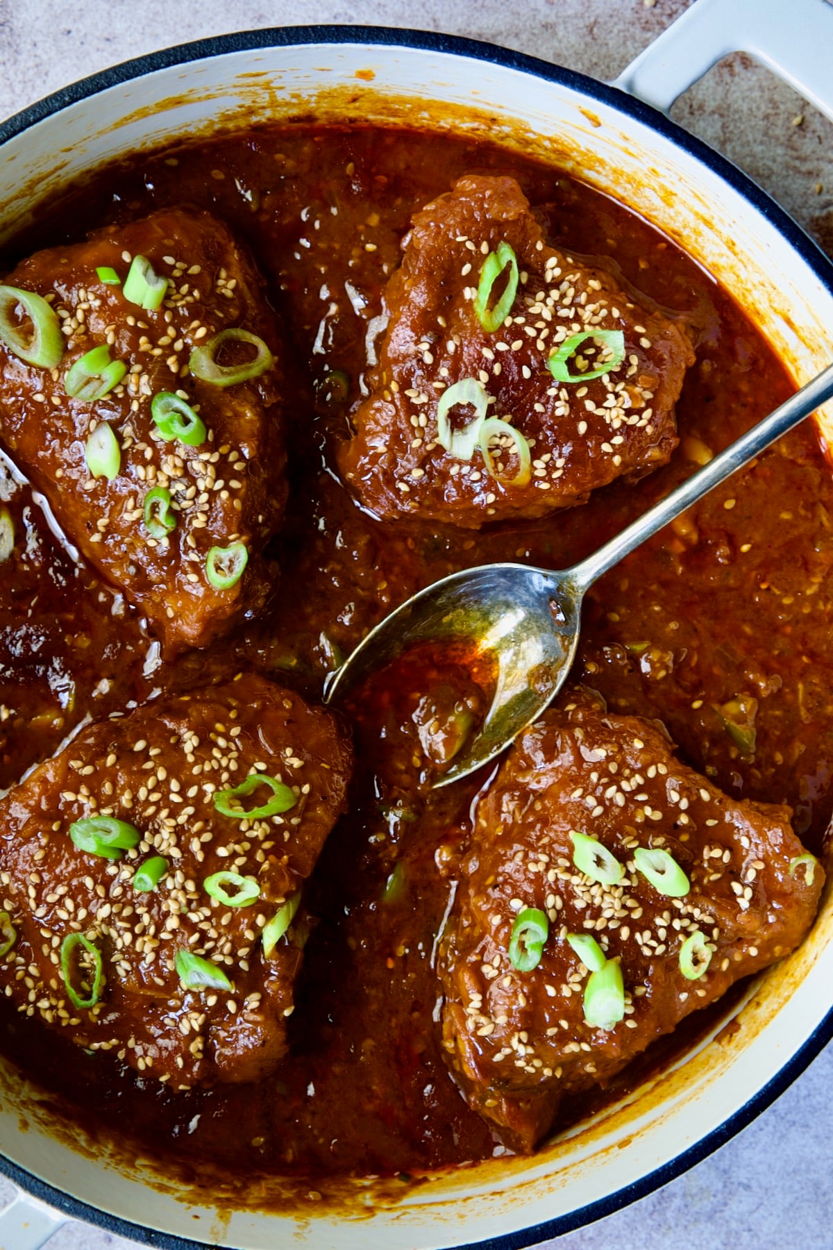 Four cooked Korean Chicken Thighs in white enamel cast iron skillet garnished with scallions and sesame seeds.