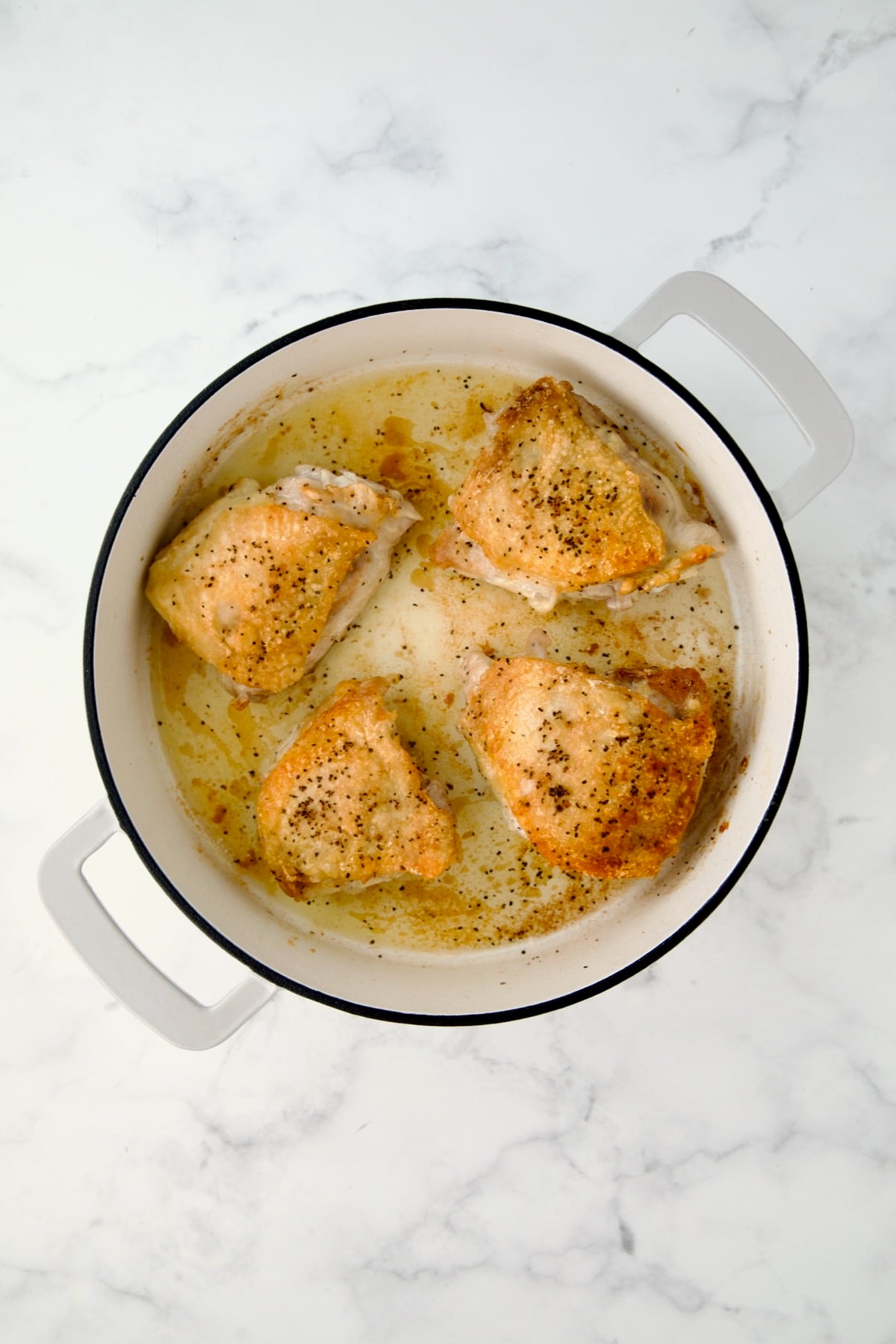 Four browned chicken thighs in white enamel cast iron skillet.
