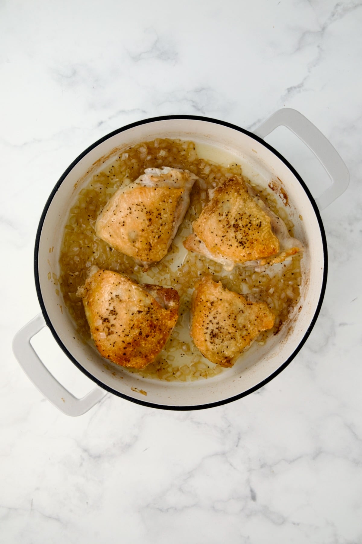 Four browned chicken thighs with onions in white enamel cast iron skillet.