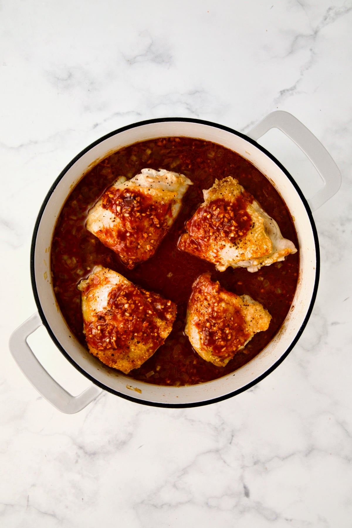 Four chicken thighs in white enamel cast iron skillet with sauce added.