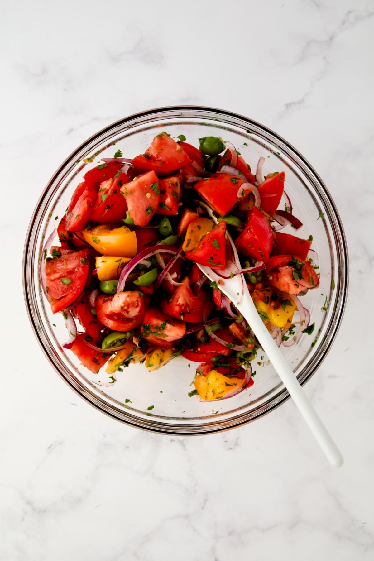 Ingredients for Panzanella in glass bowl being combined with white mixing spoon.