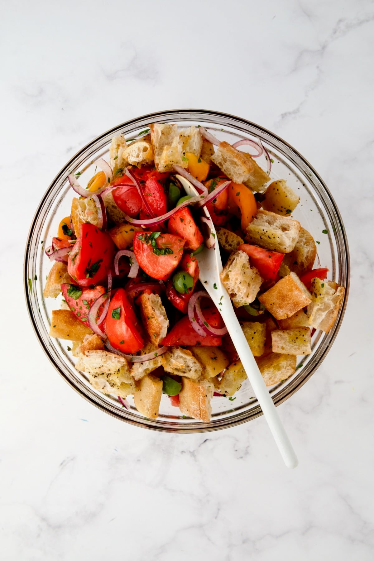 Bread added to salad ingredients being mixed with white spoon.