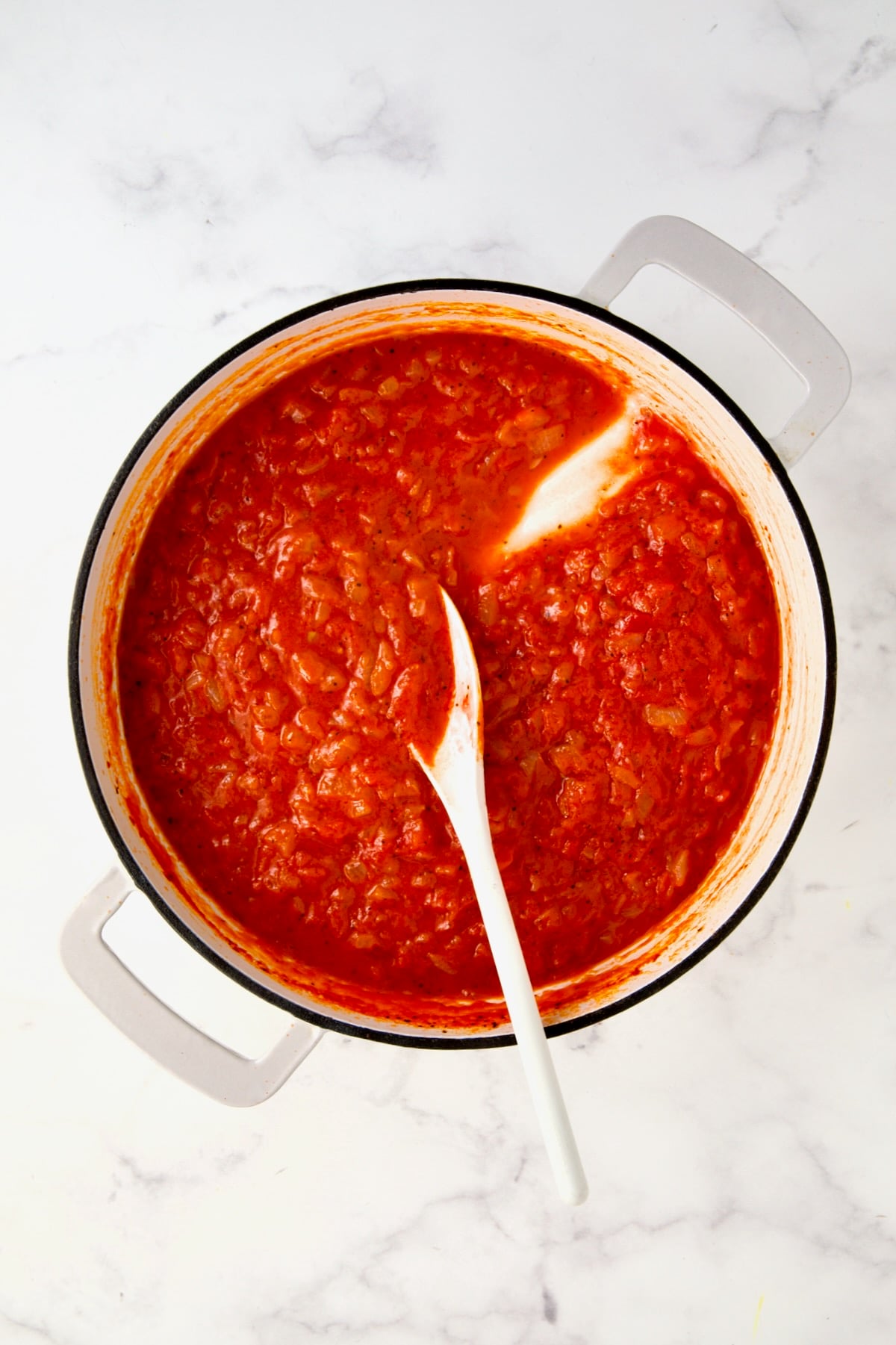 Cooked tomato sauce in white enamel cast iron skillet being stirred with white spoon.