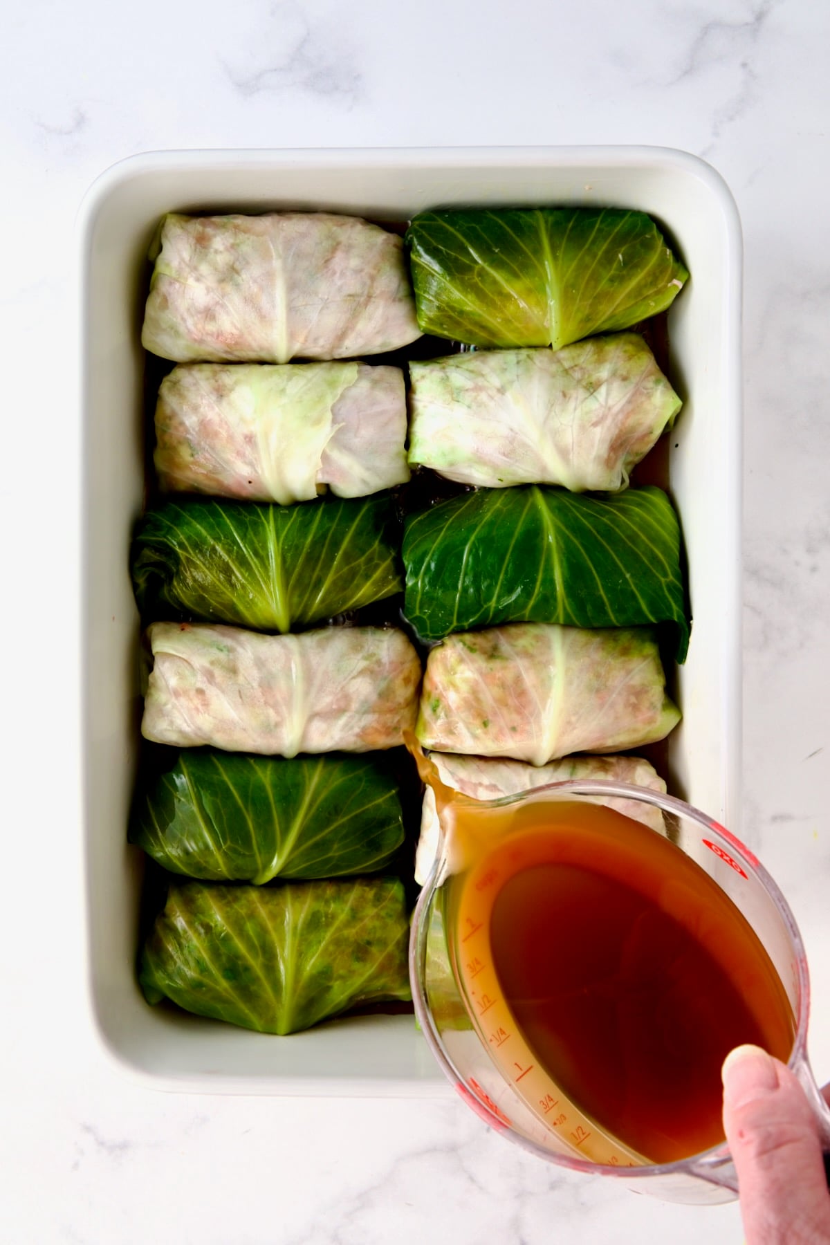 Beef broth being poured over uncooked cabbage rolls in white rectangular baking dish.