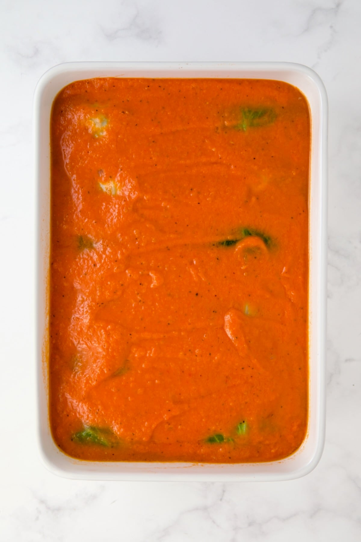Tomato sauce poured over uncooked cabbage rolls in white rectangular baking dish.