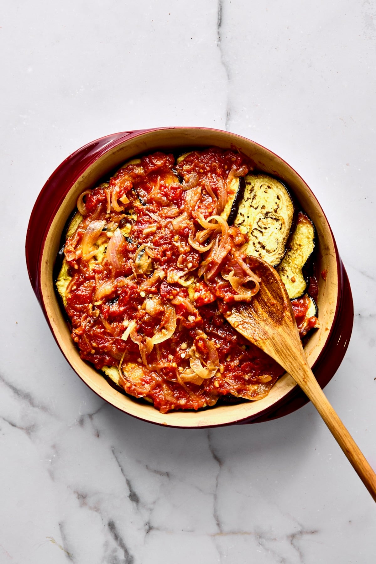 Tomato sauce being spooned over eggplant in round baking dish.