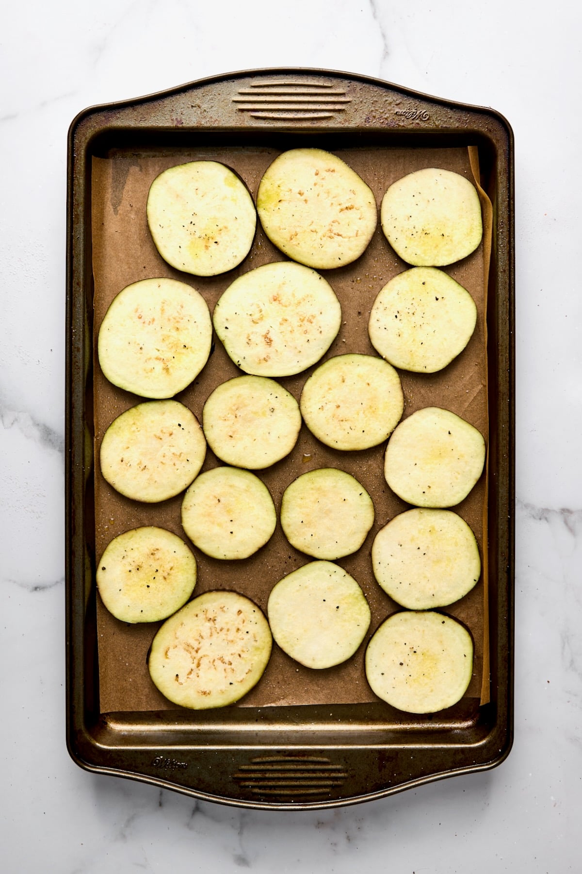Slices of eggplant on parchment paper-lined rimmed baking dish before being roasted.