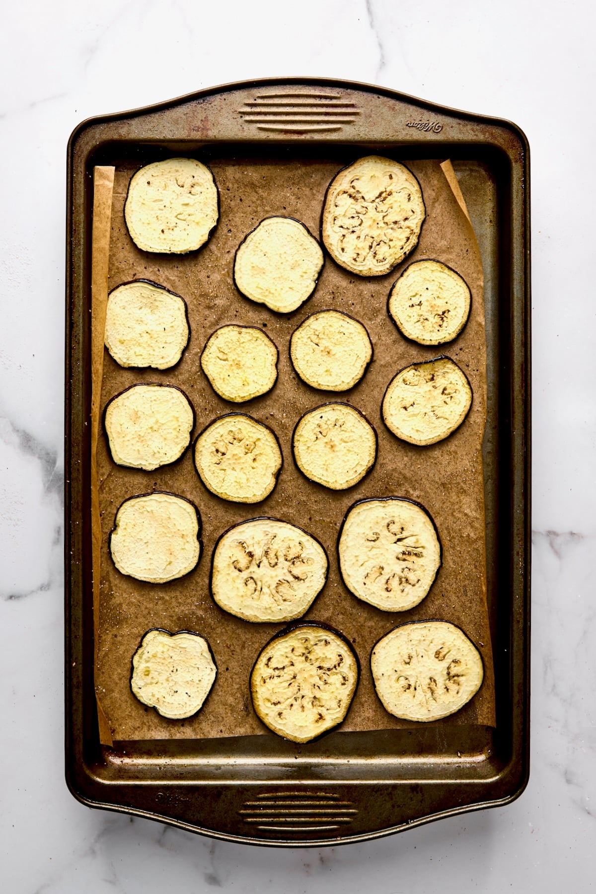 Slices of eggplant on parchment paper-lined rimmed baking dish after being roasted.