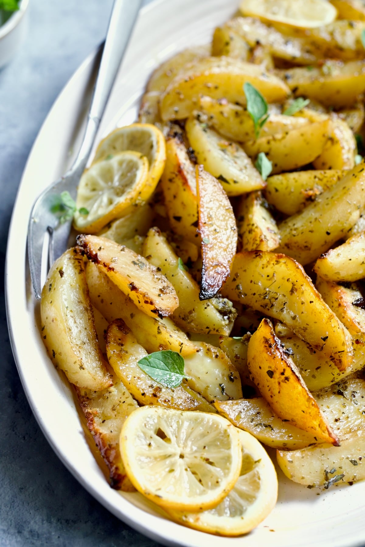 Greek Roasted Potatoes on oval white platter with serving fork garnished with lemon slices and fresh oregano leaves.