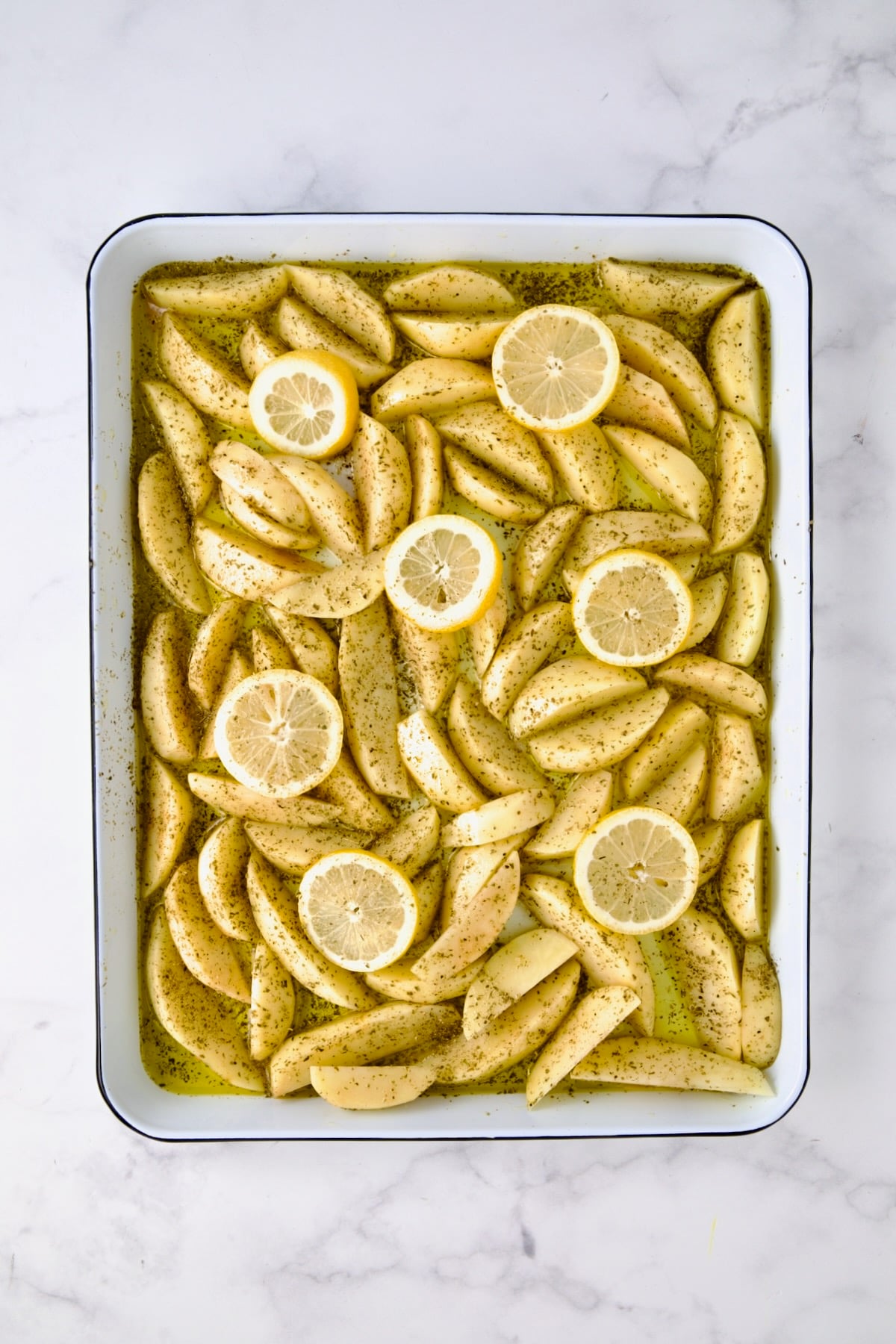 Peeled wedges of Yukon Gold potatoes on white enamel rimmed sheet pan with oregano and lemon slices.