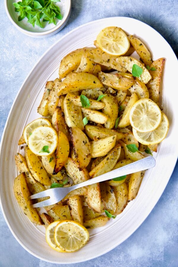 Greek Roasted Potatoes on oval white platter with serving fork garnished with lemon slices and fresh oregano leaves.