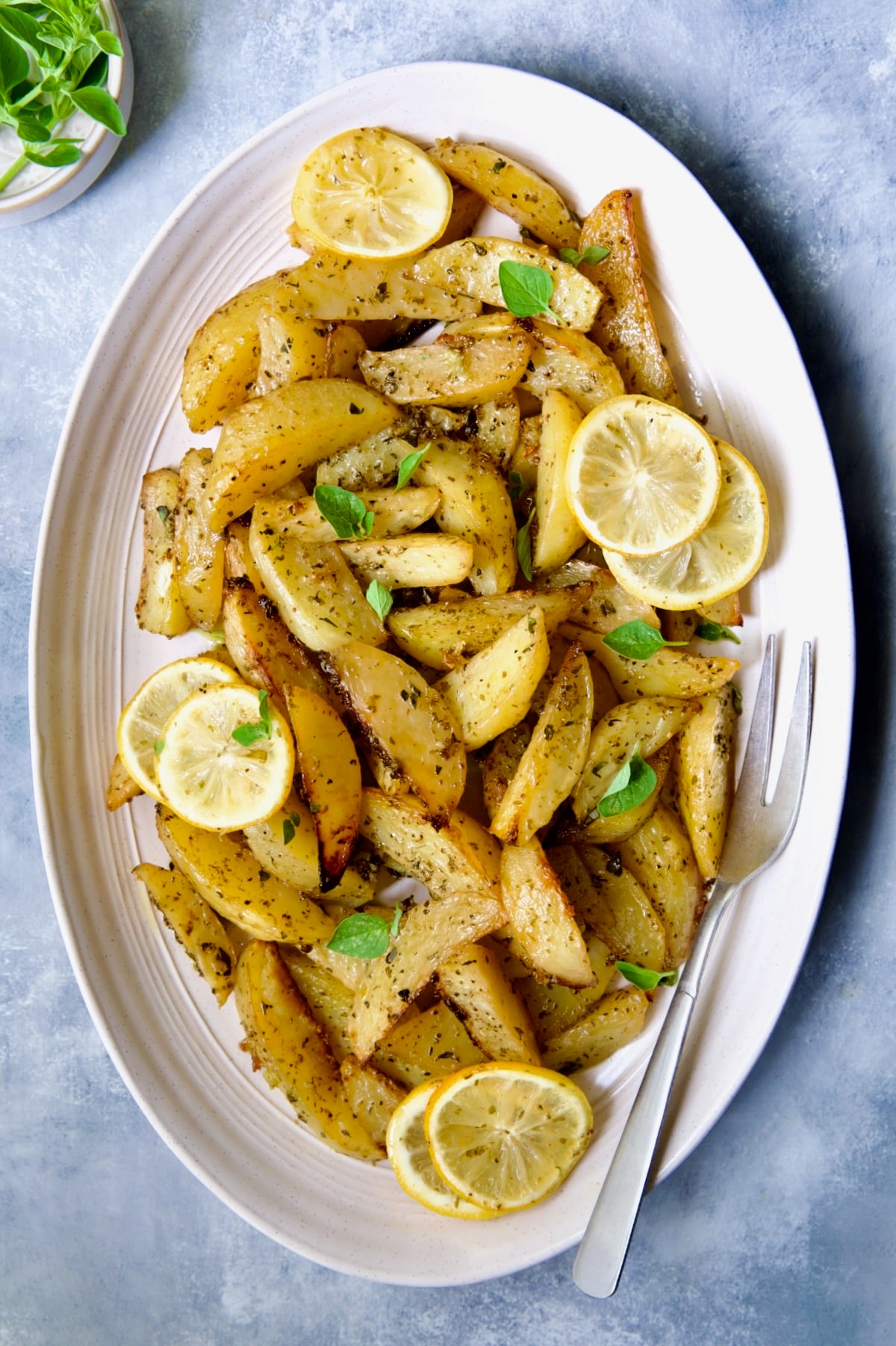 Greek Roasted Potatoes on oval white platter with serving fork garnished with lemon slices and fresh oregano leaves.