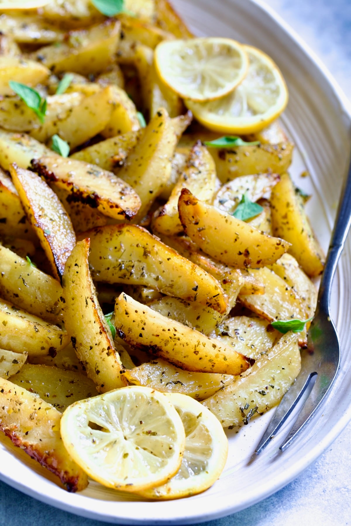 Greek Roasted Potatoes on oval white platter with serving fork garnished with lemon slices and fresh oregano leaves.
